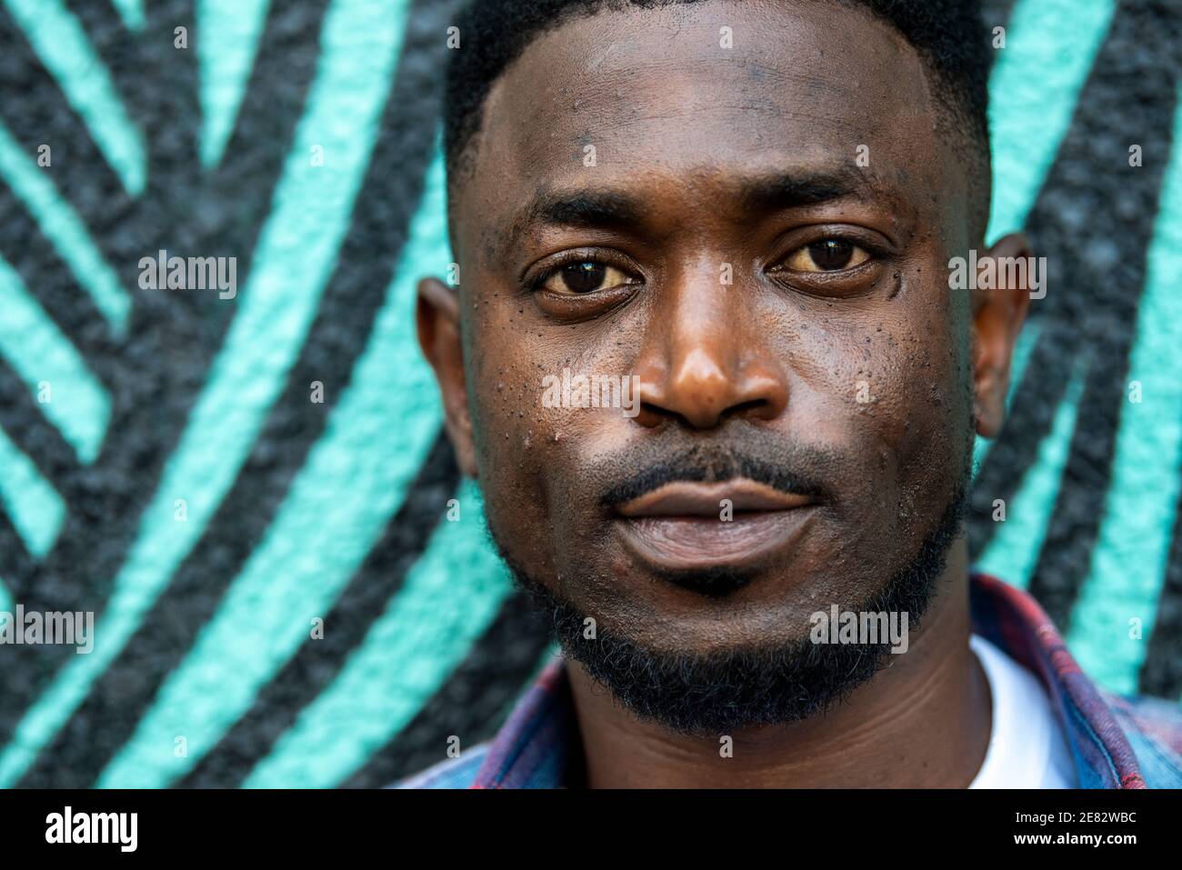 Ein Schwarzer steht vor einer türkis-schwarz bemalten Wand und blickt direkt auf die Kamera. Stockfoto