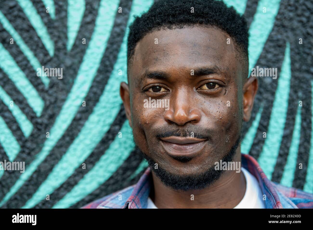Ein schwarzer Mann steht vor einer türkis-schwarz bemalten Wand und lächelt. Stockfoto