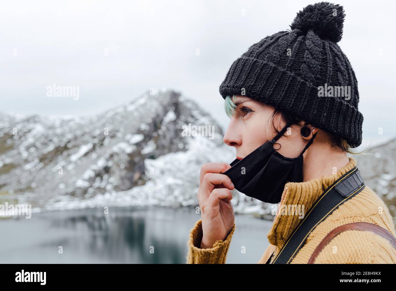 Seitenansicht ruhige junge weibliche Reisende in warmen Hut senken Gesichtsmaske und Blick weg in Kontemplation, während Sie auf Kalte Seenküste gegen maje Stockfoto