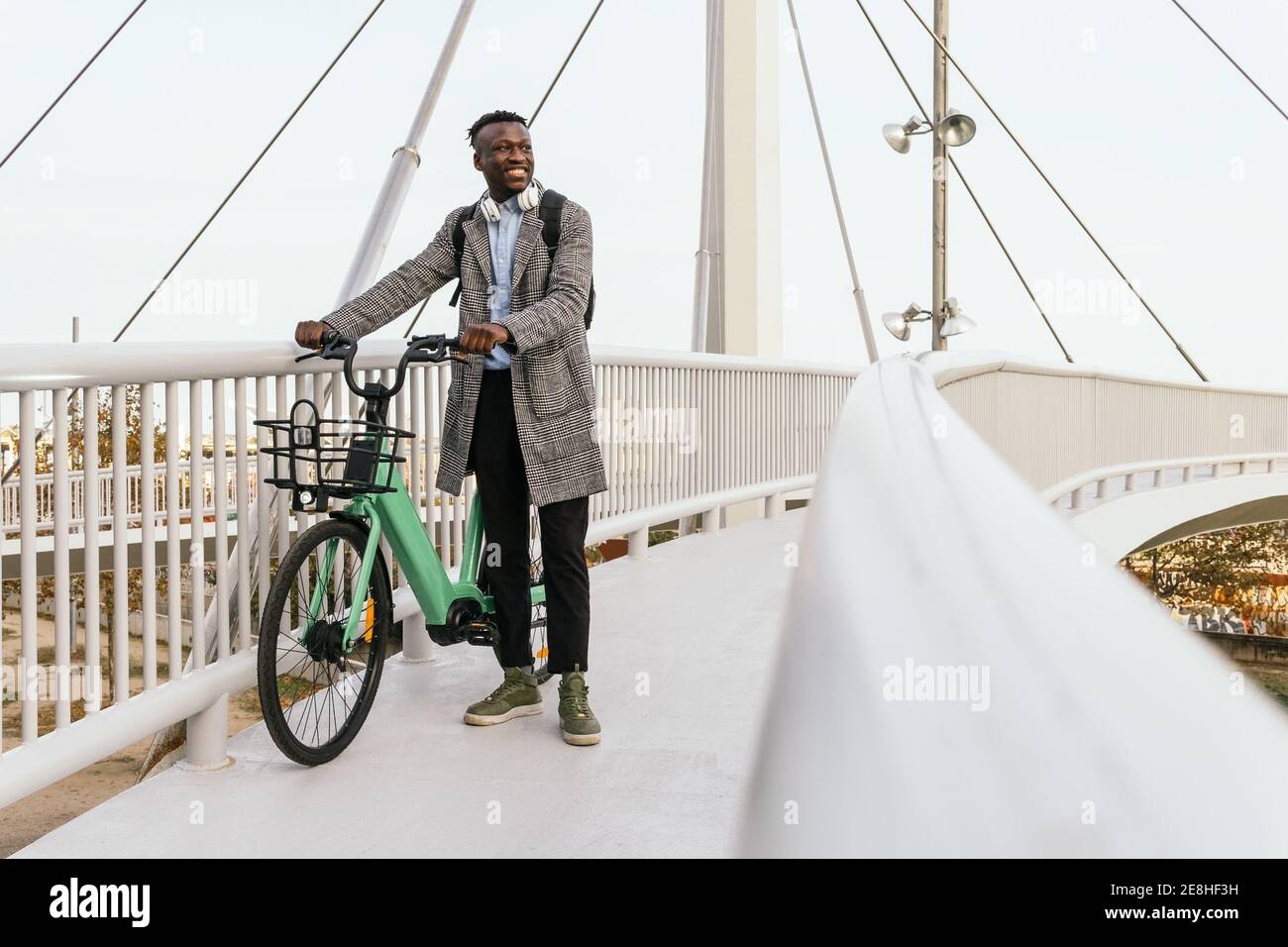 Junge lächelnde ethnische männliche Büroangestellte in formelle Outfit mit Kopfhörer und Fahrrad mit Blick auf die Hängebrücke in der Stadt Stockfoto