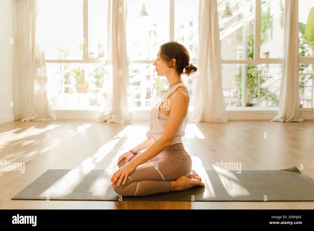 Seitenansicht einer jungen verträumten Frau in Sportkleidung, die Yoga praktiziert Mit den Armen hinter dem Rücken, während Sie mit geschlossenen Augen sitzen Sonnenlicht Stockfoto