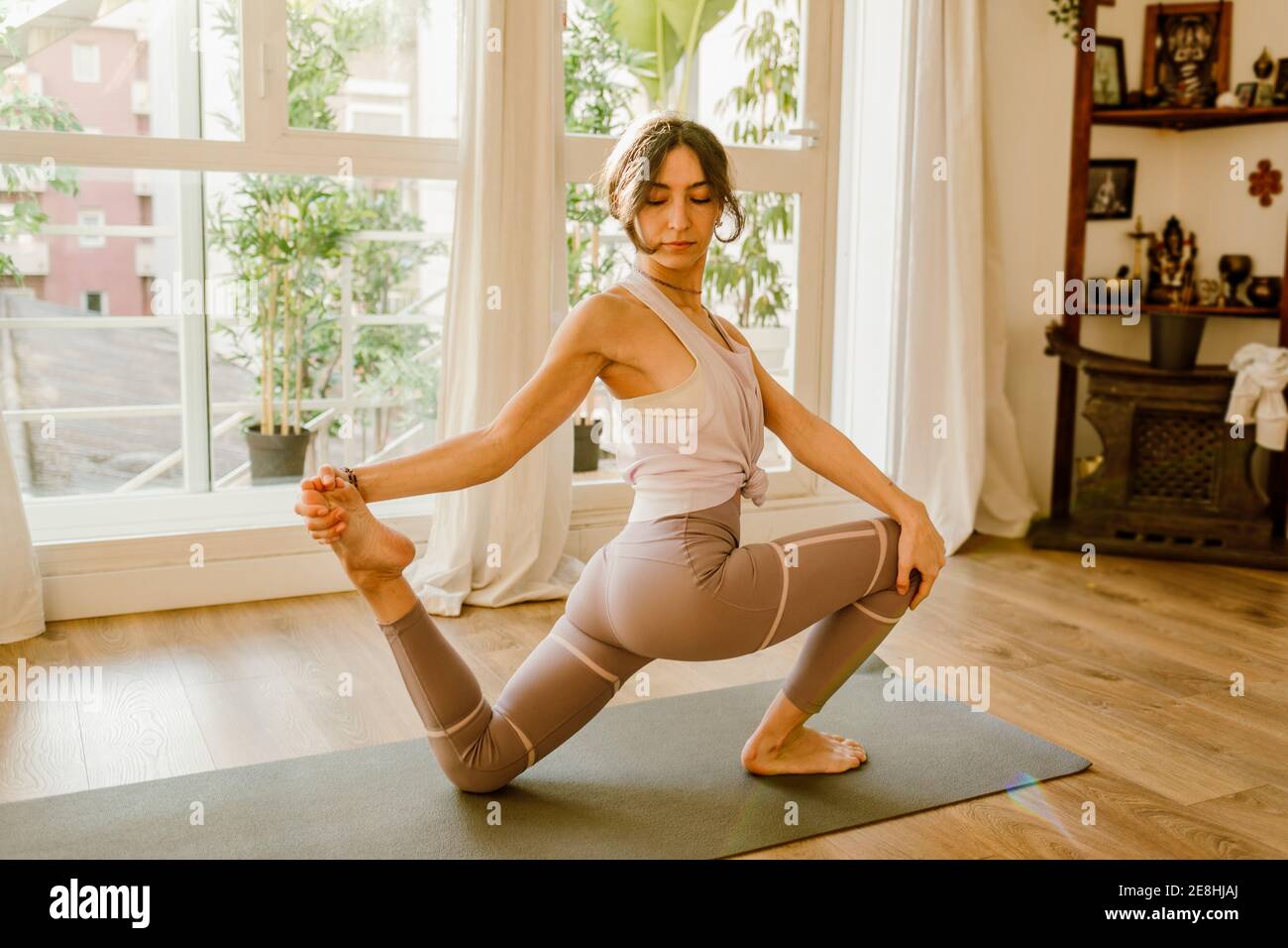 Seitenansicht der jungen flexiblen Frau in Sportkleidung stehend auf Knie und Fuß berühren, während Yoga im Haus praktiziert wird Stockfoto