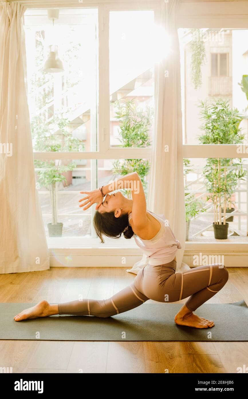 Seitenansicht der jungen flexiblen Frau in Sportkleidung stehend auf Knie und Fuß berühren, während Yoga im Haus praktiziert wird Stockfoto