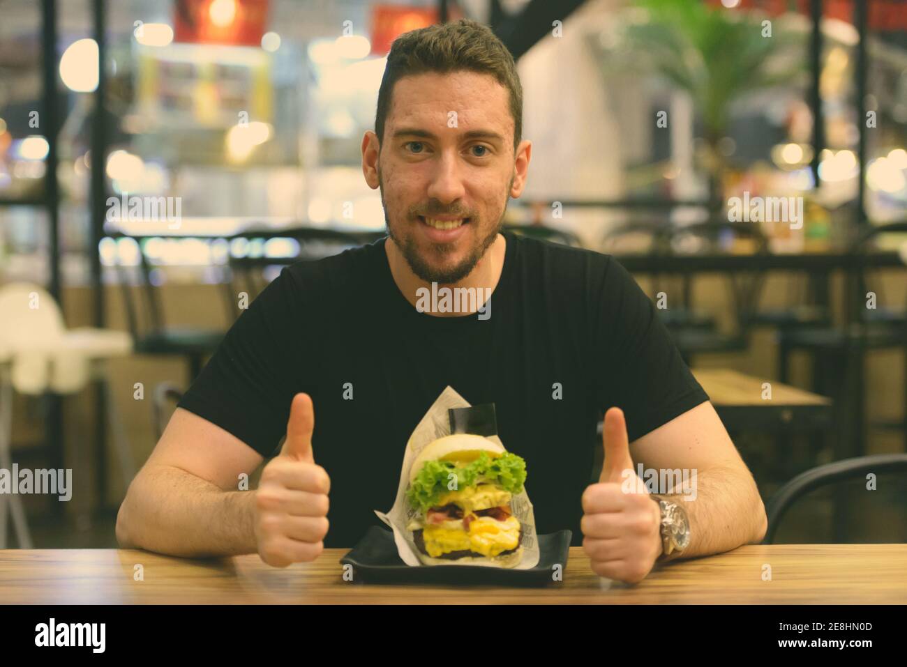 Glücklicher Kaukasischer Mann, der lächelt und im Restaurant Daumen nach oben gibt Serviert mit leckeren Burger Stockfoto