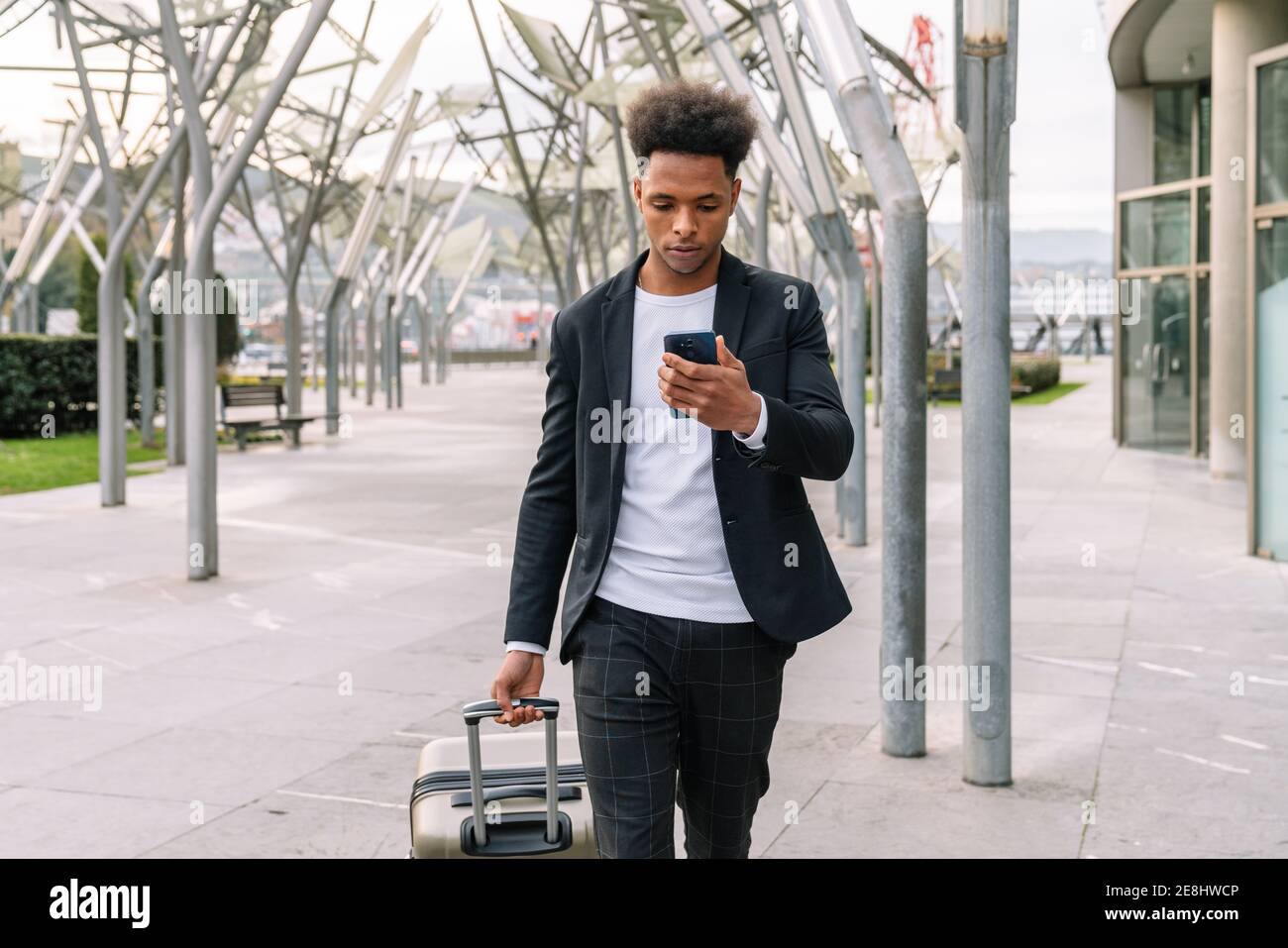 Serious African American männlichen Touristen zu Fuß mit Koffer im Flughafen Und die Abreisezeit auf dem Smartphone überprüfen Stockfoto