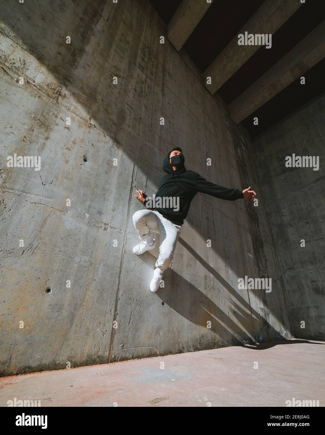 Von unten unerkennbarer junger Rüde im trendigen Outfit und Maske mit Trick an Betonwand beim Tanzen in Abandoned Gebäude an sonnigen Tag Stockfoto