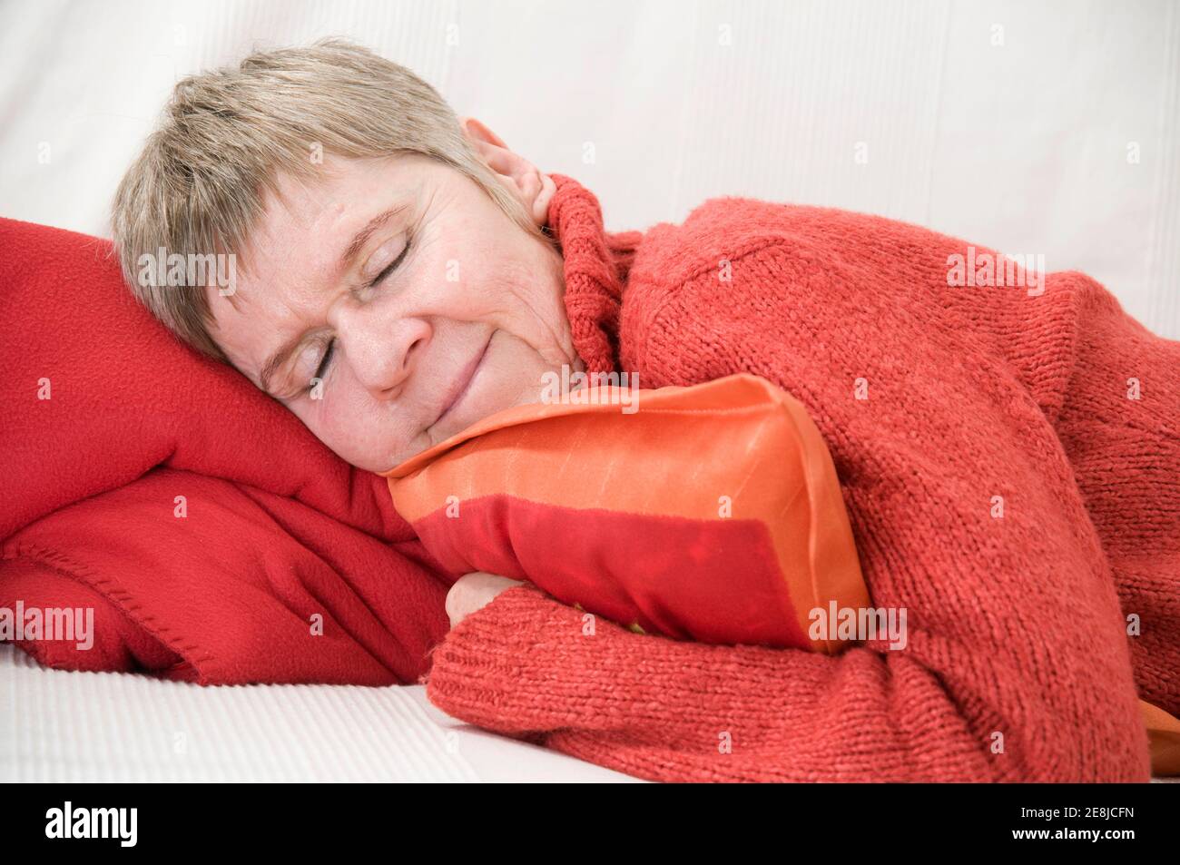 Oberkörperansicht einer älteren Frau, die seitwärts auf einer weißen Couch liegt, mit geschlossenen Augen und einem Kissen in den Armen. Stockfoto