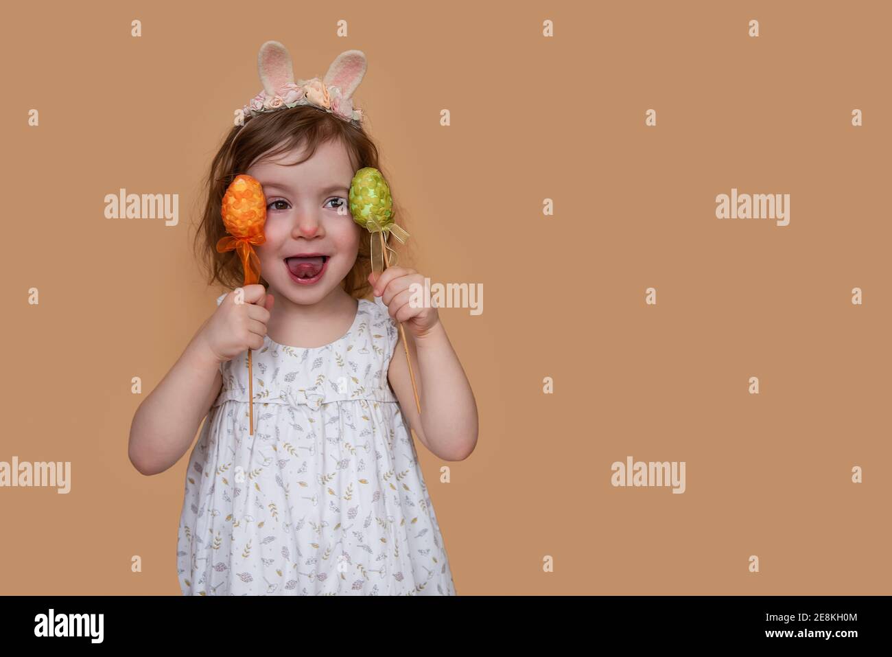 Positives Porträt eines kleinen Mädchens mit Kaninchenohren auf dem Kopf, zeigt ihre Zunge, die ein orangegrünes Ei hält. Isolierter hellorangefarbener Hintergrund, kopieren Stockfoto