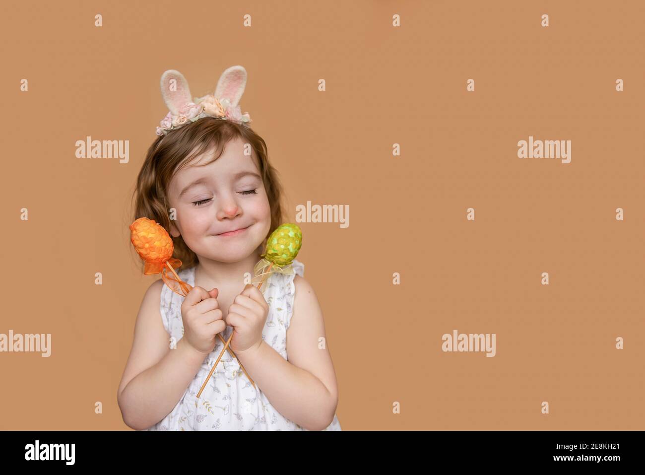 Positives Porträt eines kleinen Mädchens mit Kaninchenohren auf dem Kopf, Träume von Schokolade, in ihren Händen ein orange grünes Ei. Isolierter hellorangefarbener Hintergrund Stockfoto