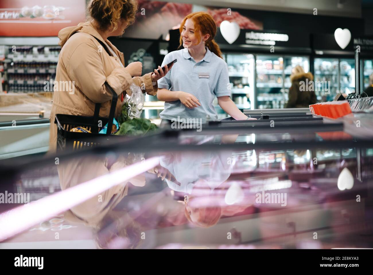 Weibliche Kundin, die eine Supermarktarbeiterin um Hilfe bei der Suche nach einem Lebensmittel bittet. Frau hält ihr Handy und bittet um Hilfe zu einem weiblichen Lebensmittelgeschäft e Stockfoto