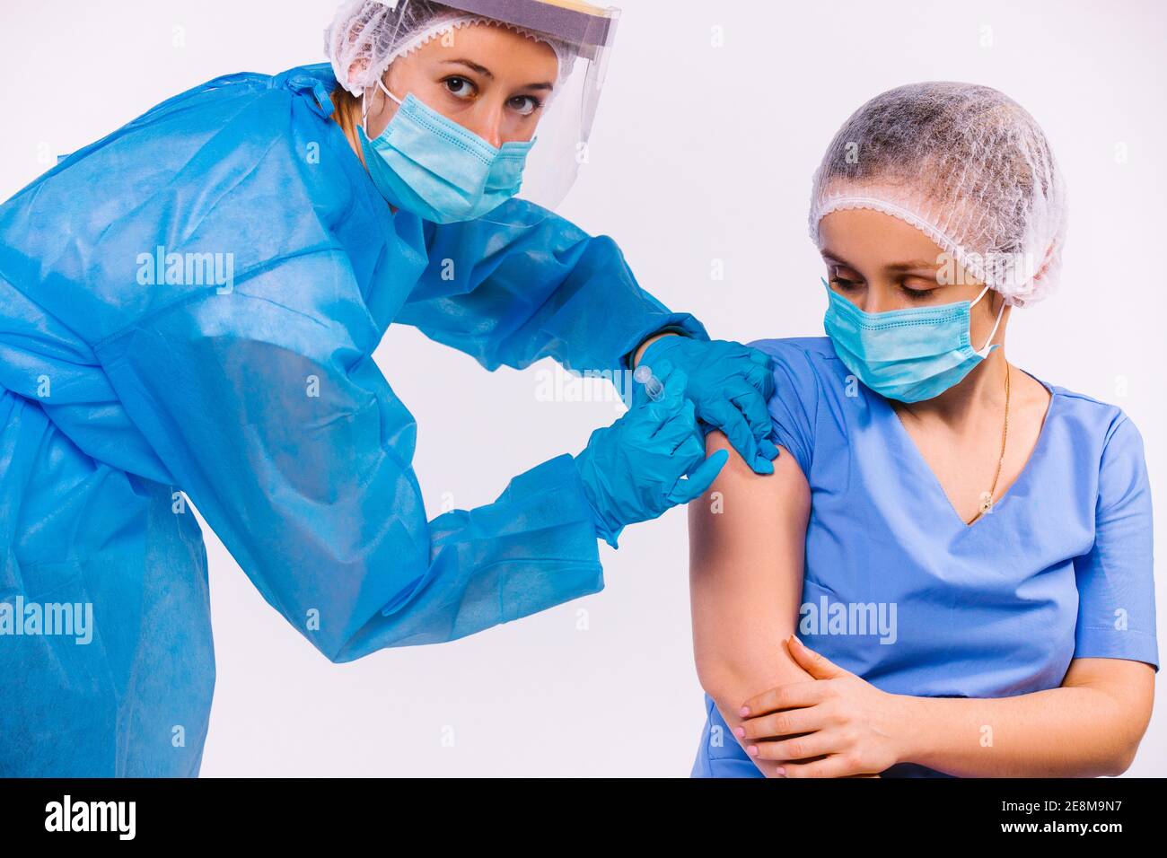 Eine Krankenschwester in Schutzausrüstung, in einer medizinischen Maske, macht eine junge Ärztin einen Impfstoff gegen covid 19 und schaut auf die Kamera. Weißer Hintergrund. Stockfoto