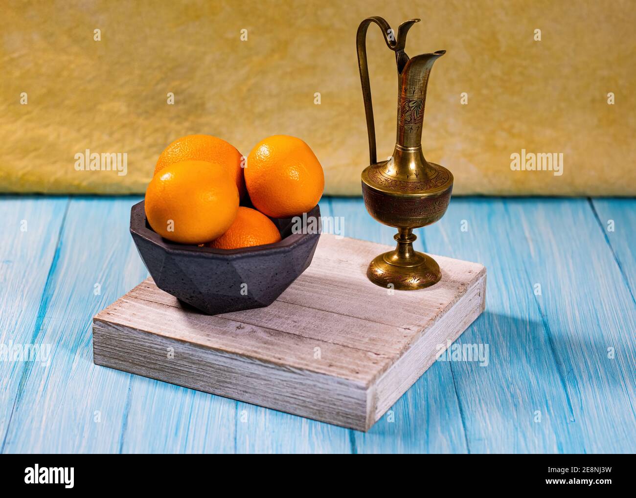 Eine Gruppe von Orangen in einer geodätischen Betonschale und eine geätzte Messingurne auf einer cyanfarbenen Basis mit hellgelb-oranger Kulisse. Stockfoto