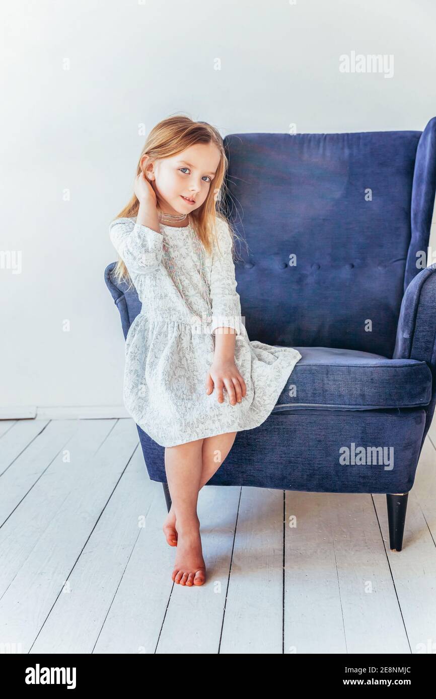 Süße kleine Mädchen in weißem Kleid sitzt auf modernen gemütlichen blauen Stuhl entspannen in weißen hellen Wohnzimmer zu Hause drinnen. Kindheit Schulkinder out Stockfoto