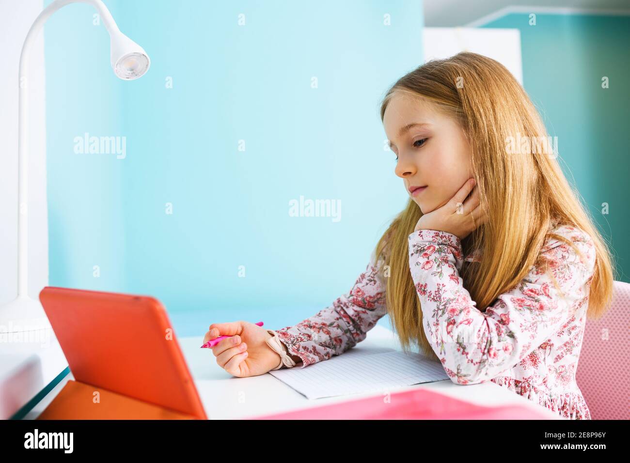 Alter der Grundschule des Kindes Sitzen Sie am Schreibtisch mit leerem Übungsheft, halten Sie den Stift und schauen Sie auf den Tablet-Bildschirm, lernen Sie zu Hause während des Lockdown Stockfoto