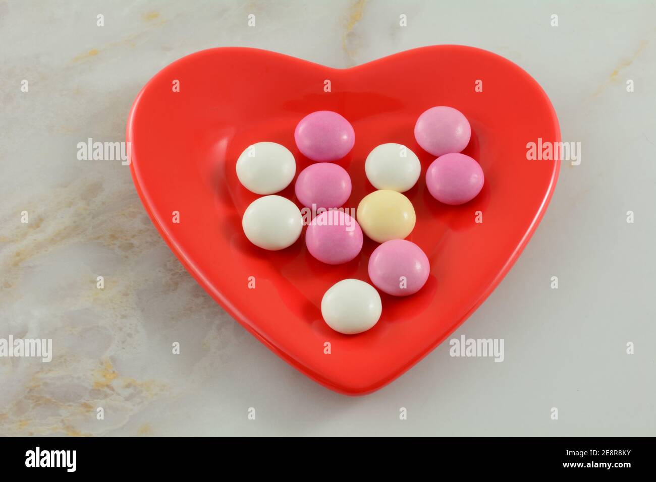 Rosa, weiß und gelb Valentinstag Süßigkeiten bedeckt Schokoladenstücke auf roten herzförmigen Teller auf Tisch Stockfoto