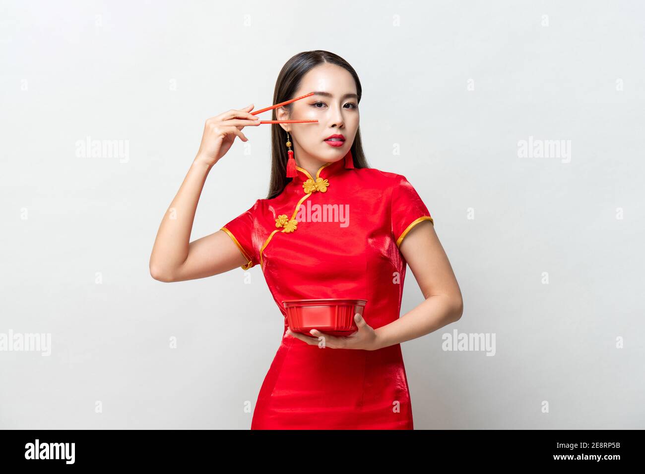 Schöne asiatische Frau in traditionellen roten chinesischen Qipao Kleid suchen An der Kamera halten Lebensmittelbox und Essstäbchen im Studio isoliert Grauer Hintergrund Stockfoto