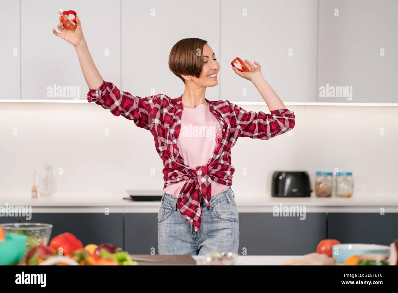 Nahaufnahme. Kurze Haare Frau im roten Hemd spielen mit Essen, schneiden Scheiben von süßem Papier beim Kochen in der weißen Küche. Gesunde Ernährung zu Hause. Gesund Stockfoto