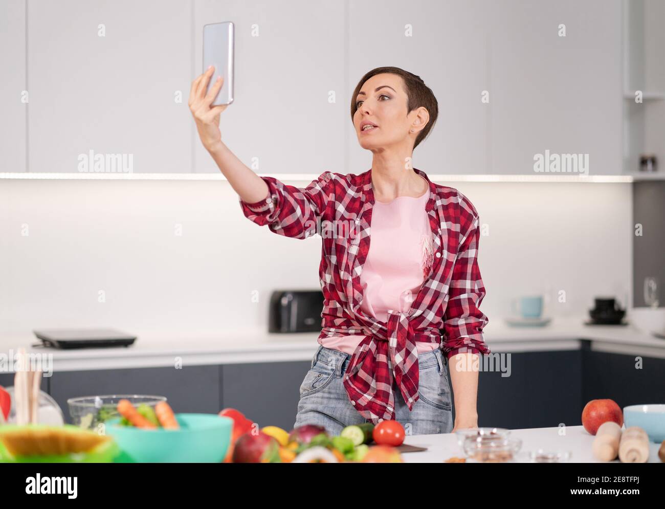 Hübsche Frau glücklich, Selfie mit ihrem Smartphone in ihrem neuen Zuhause beim Kochen frischen Salat trägt ein kariertes Hemd mit einem bob Frisur. Gesund Stockfoto