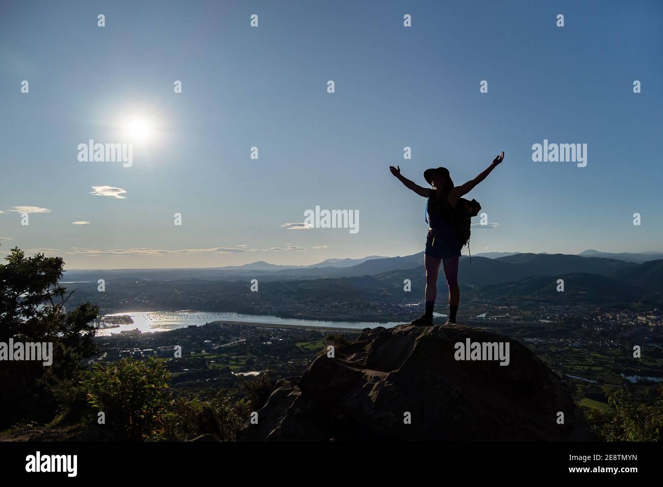 Silhouette des Mädchens in Hut, Flecken auf einem Felsen und breitet ihre Hände an den Seiten. Eine Stadt, Berge, Meer im Hintergrund Stockfoto