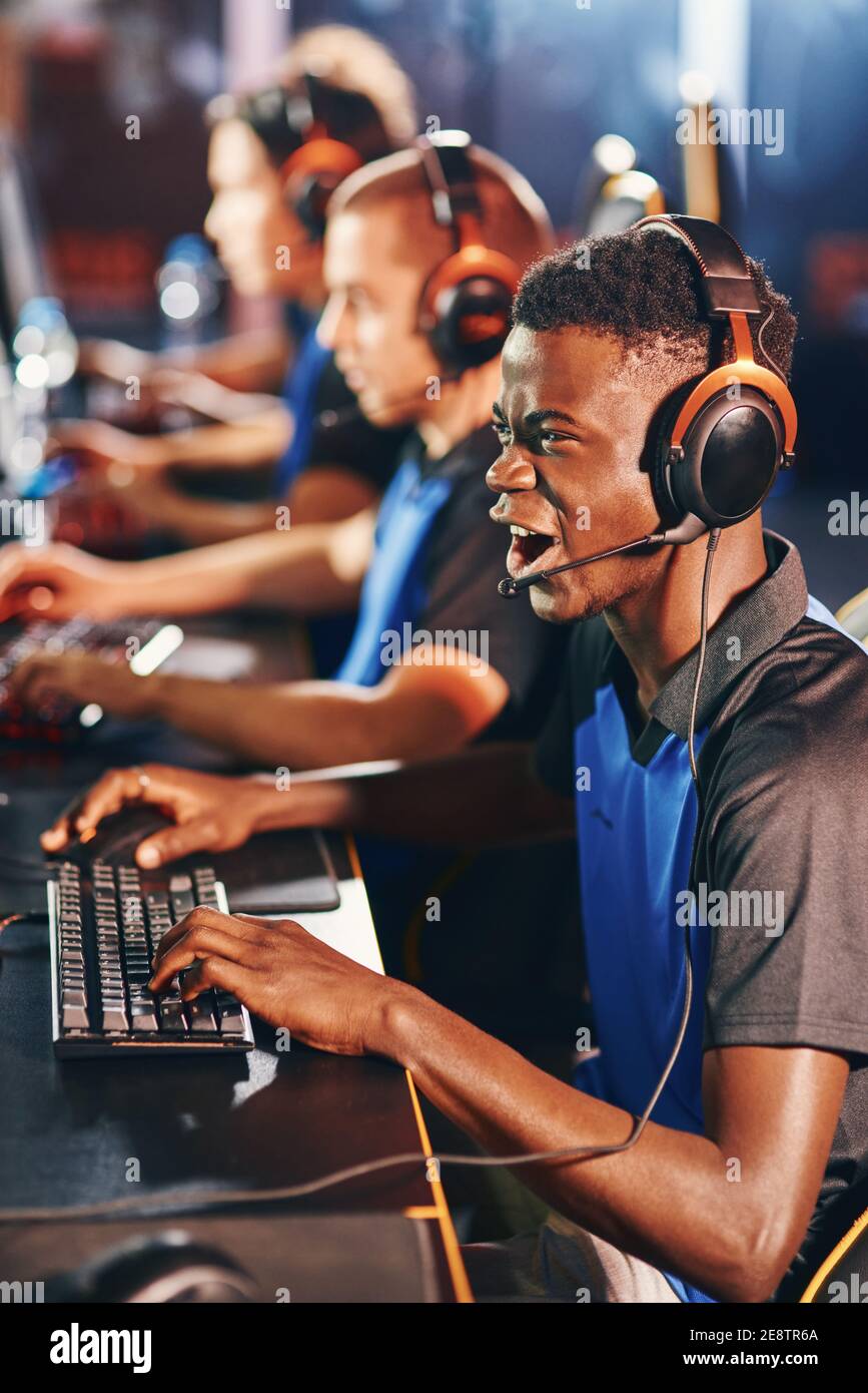 Genießen. Seitenansicht eines jungen aufgeregt afrikanischen Kerl, männliche Cybersport-Gamer mit Kopfhörern spielen Online-Videospiele, während die Teilnahme an eSport-Turnier. Videospiele Online-Wettbewerb Stockfoto