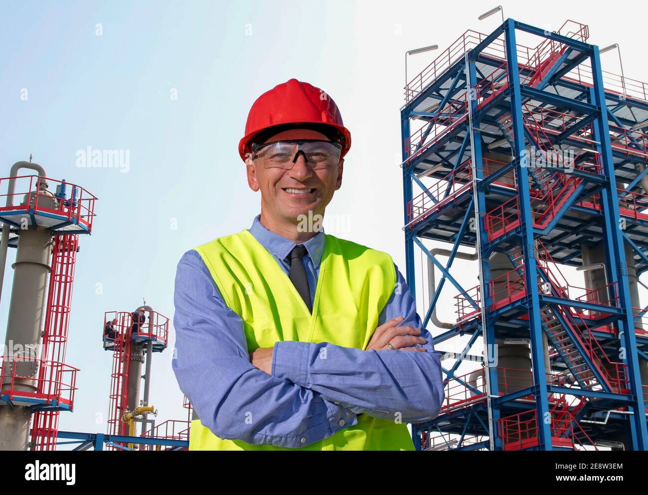 Happy Manager in Red Hardhat, Blue Shirt und Yellow Vest vor der Öl- und Gasraffinerie. Porträt von Vorarbeitern, Vorgesetzten oder Geschäftsleuten Stockfoto