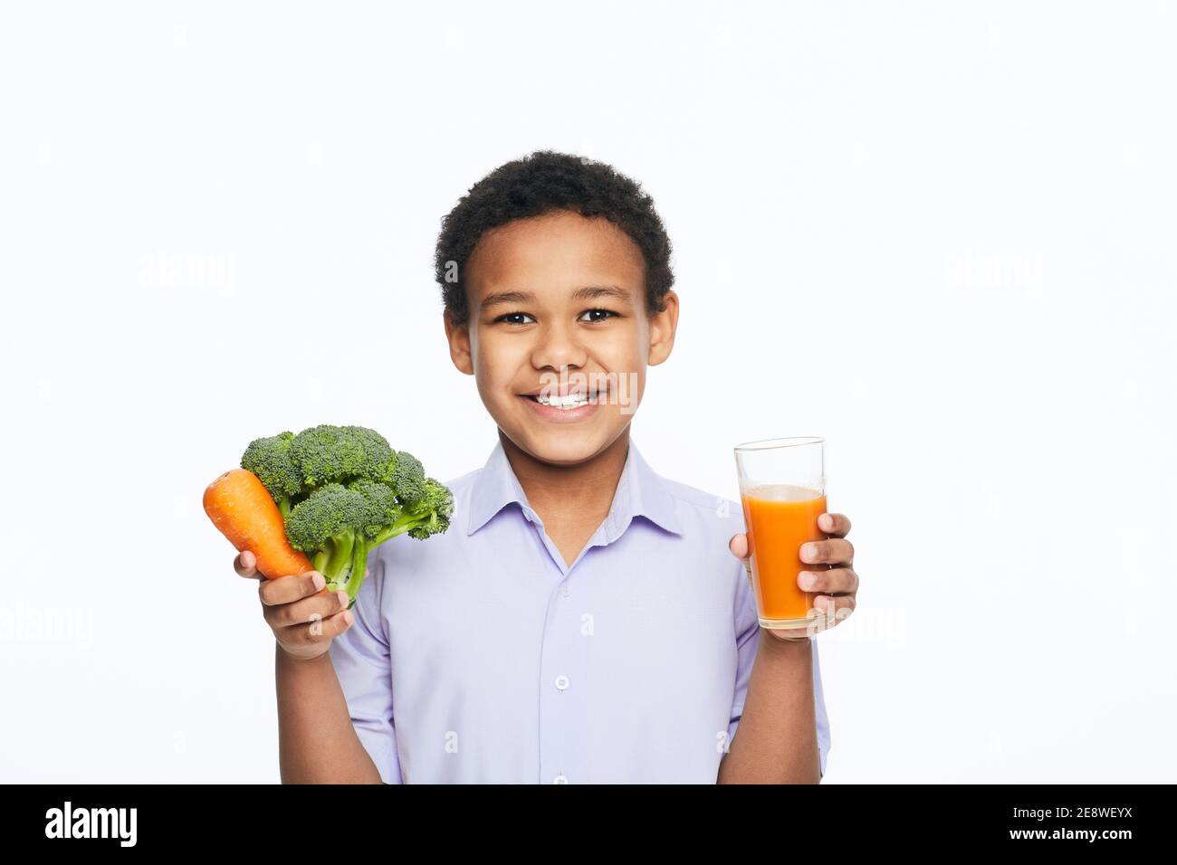 Der afroamerikanische Junge hält Karotten, Brokkoli und Karottensaft auf weißem Hintergrund. Gesund essen für Kinder Stockfoto