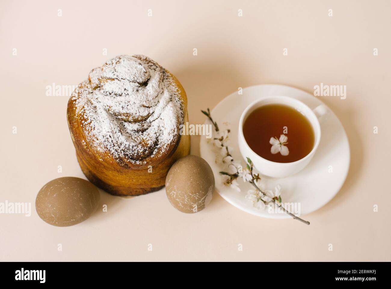 Ein traditioneller Osterkuchen, bestreut mit Puderzucker, beigefarbenen Eiern und einer Tasse schwarzen Tee in einem weißen Becher und einem Zweig Apfelblüte. Holida Stockfoto