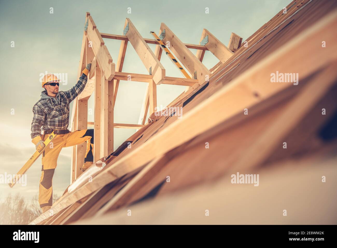 Thema Bauindustrie. Kaukasischen Auftragnehmer Arbeiter in seinen 40er Jahren mit Sprit Level Tool in der Hand auf dem Dach des neu gebauten Holzhaus Dach Str Stockfoto