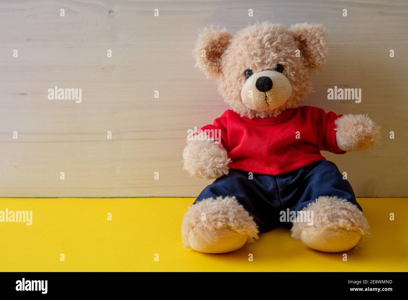 Teddybär sitzt in einem leeren Raum, gelber Boden, weißer Holzwandhintergrund, Kind allein trägt rotes T-Shirt im Kinderzimmer Stockfoto