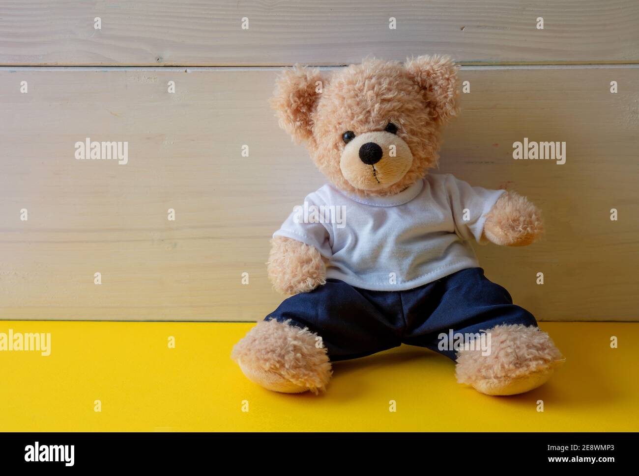 Teddybär sitzt in einem leeren Raum, gelber Boden, weißer Holzwandhintergrund, Kind allein trägt weißes T-Shirt im Kinderzimmer Stockfoto