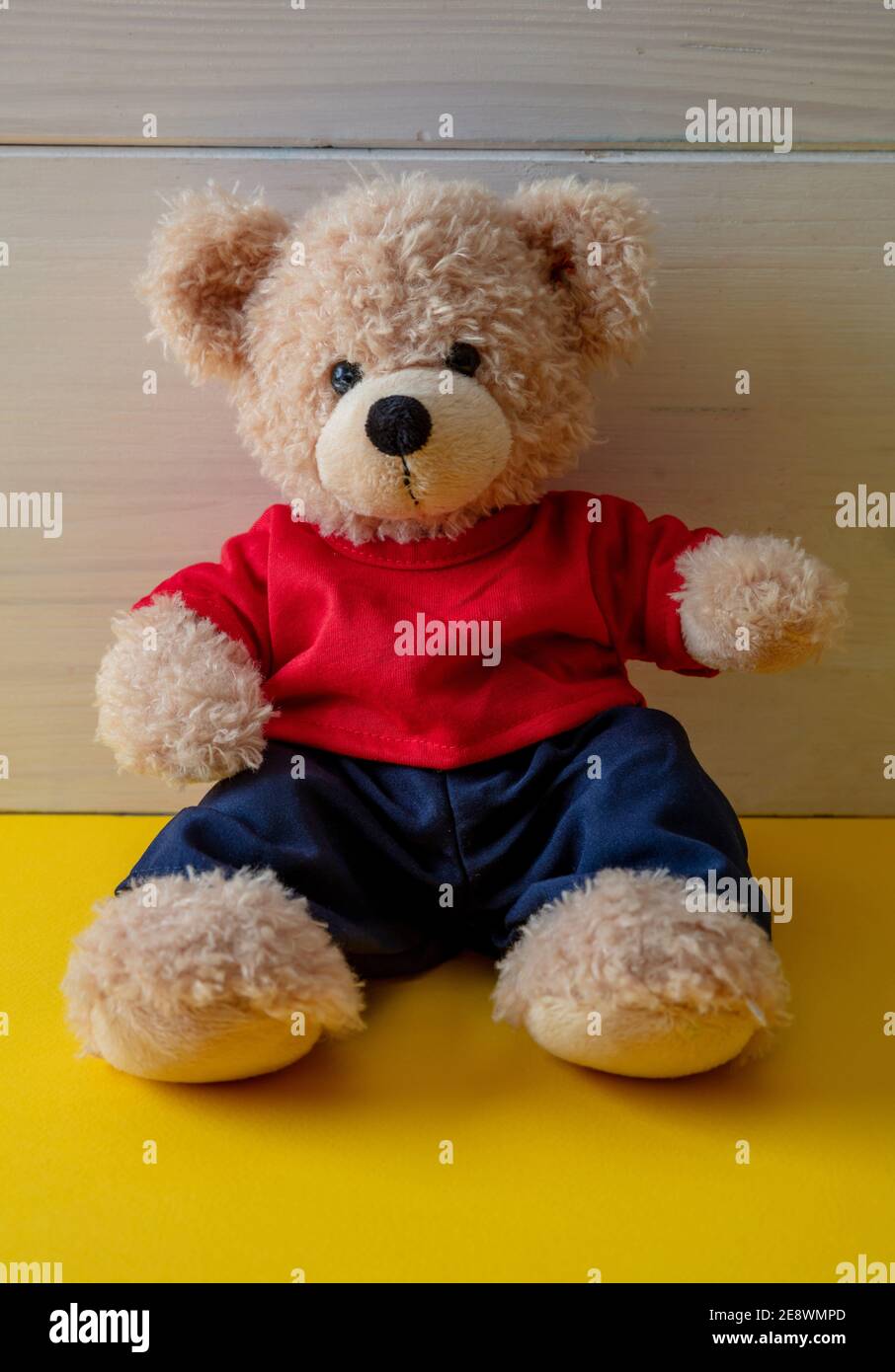 Teddybär sitzt in einem leeren Raum, gelber Boden, weißer Holzwandhintergrund, Kind allein trägt rotes T-Shirt im Kinderzimmer Stockfoto
