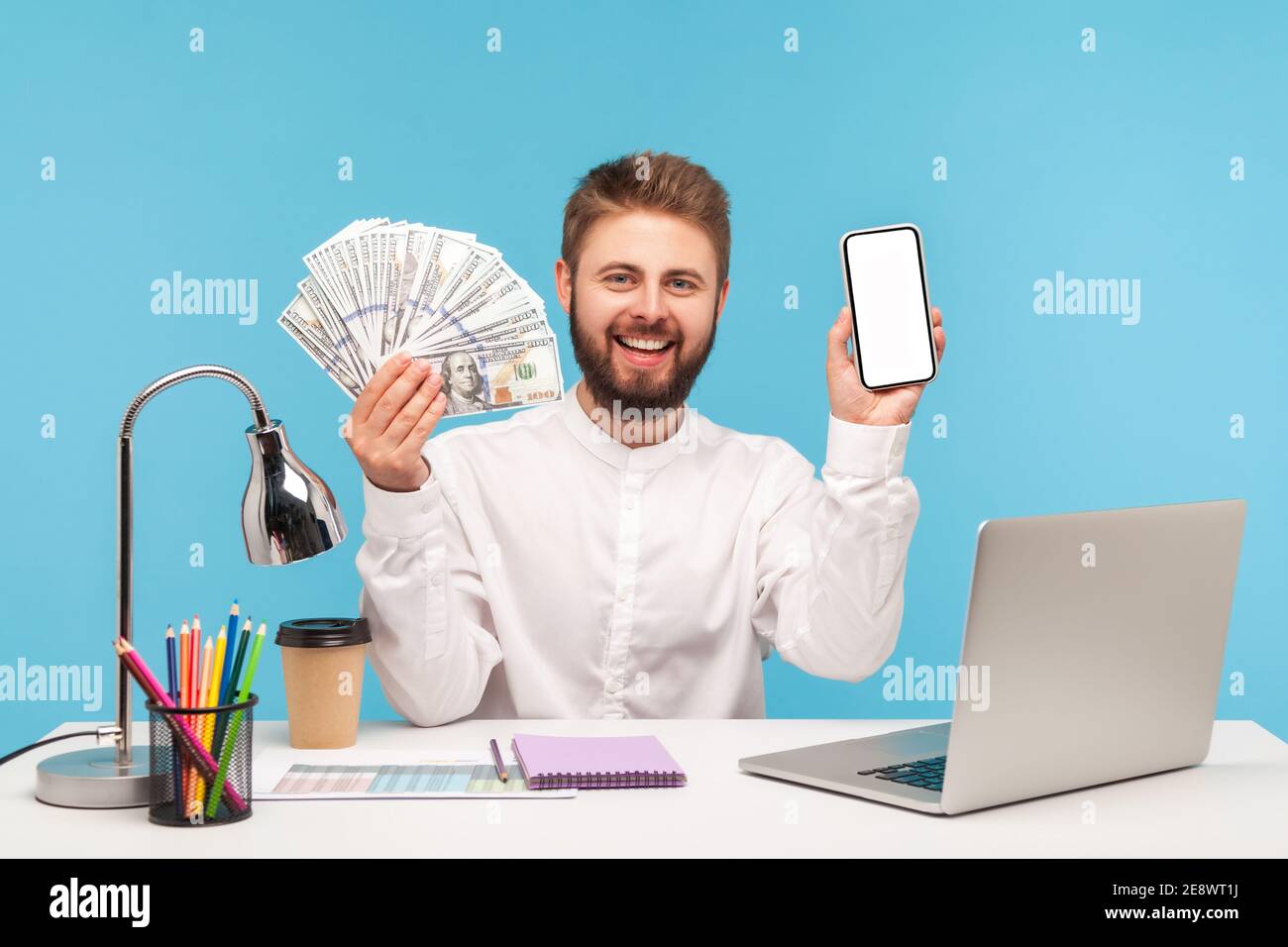 Positiv lächelnder Mann mit Bart in weißem Hemd zeigt viele hundert Dollar Scheine und weißen Bildschirm Smartphone, Banking und Gutschrift in Online-App. ICH Stockfoto