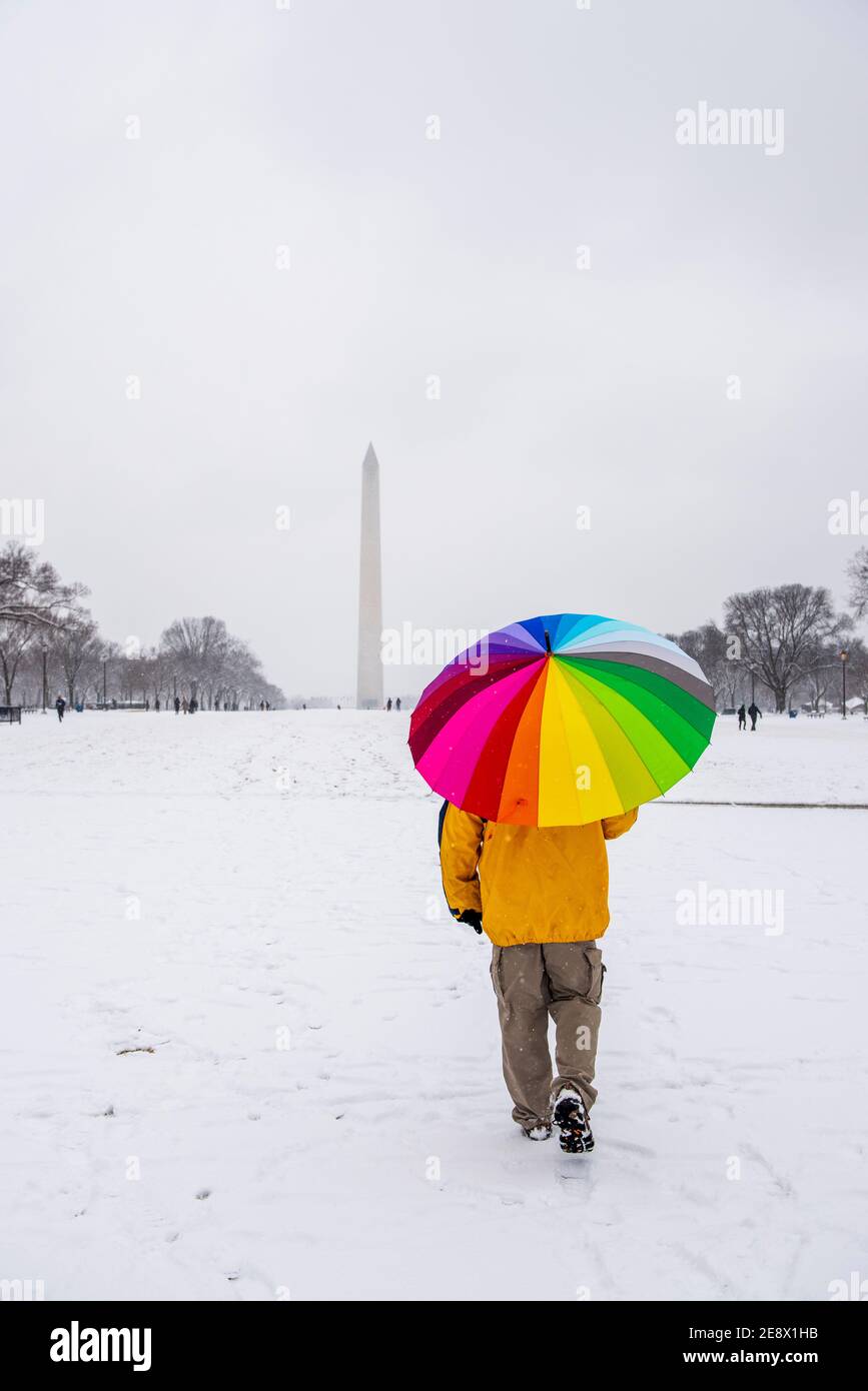Ein Mann trägt einen bunten Regenschirm während eines verschneiten Tages auf der National Mall in Washington, D.C. das Washignton Monument kann in der Ferne gesehen werden. Stockfoto