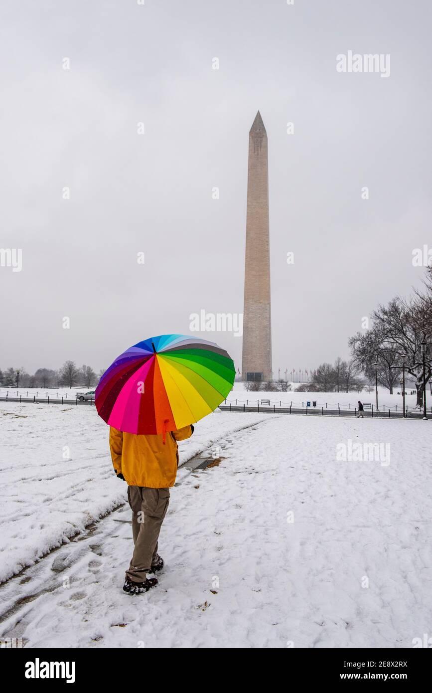 Ein Mann trägt einen bunten Regenschirm während eines verschneiten Tages auf der National Mall in Washington, D.C. das Washignton Monument kann in der Ferne gesehen werden. Stockfoto