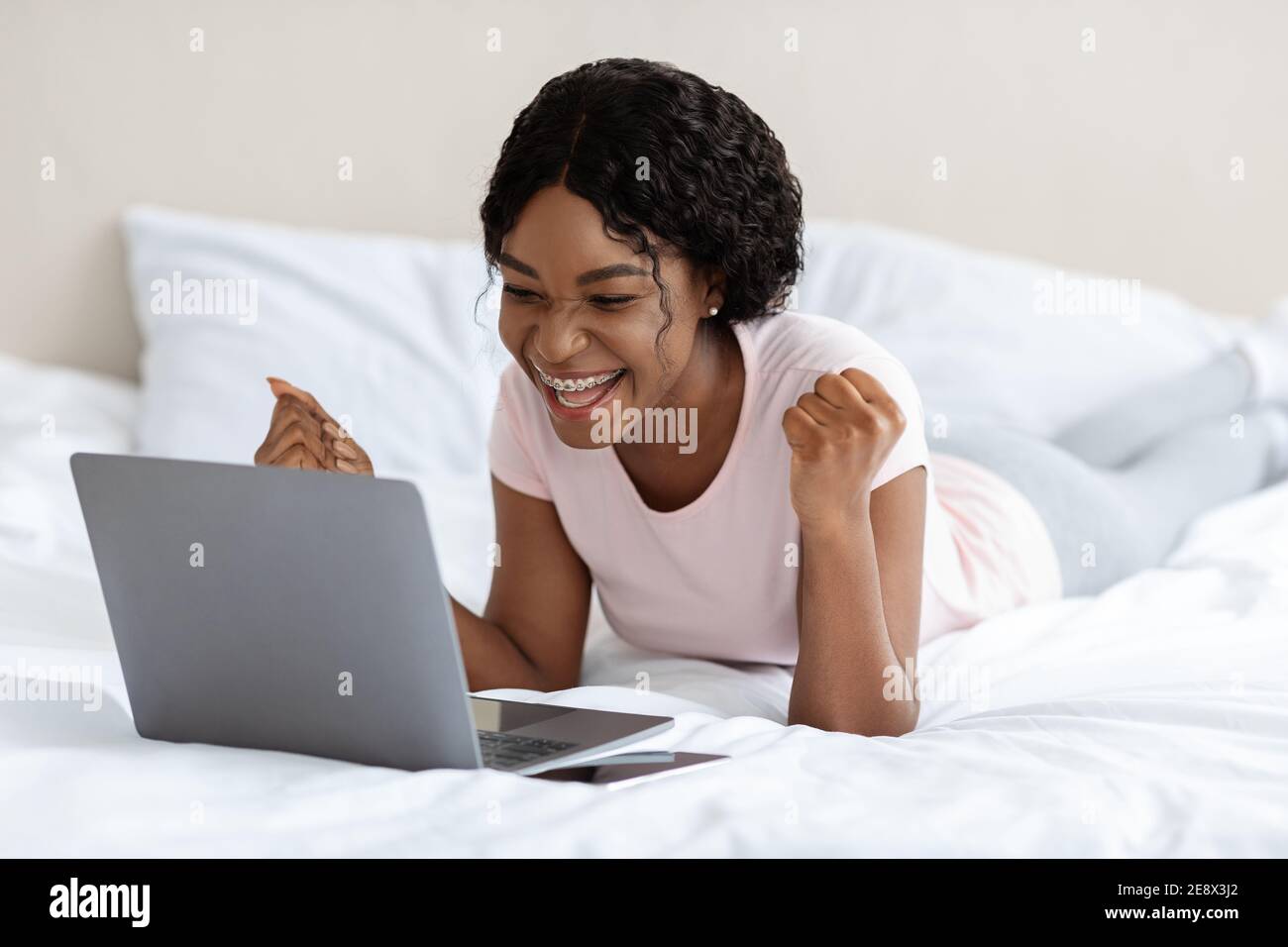 Positive afrikanische Frau feiert Erfolg, lag mit Laptop auf dem Bett Stockfoto