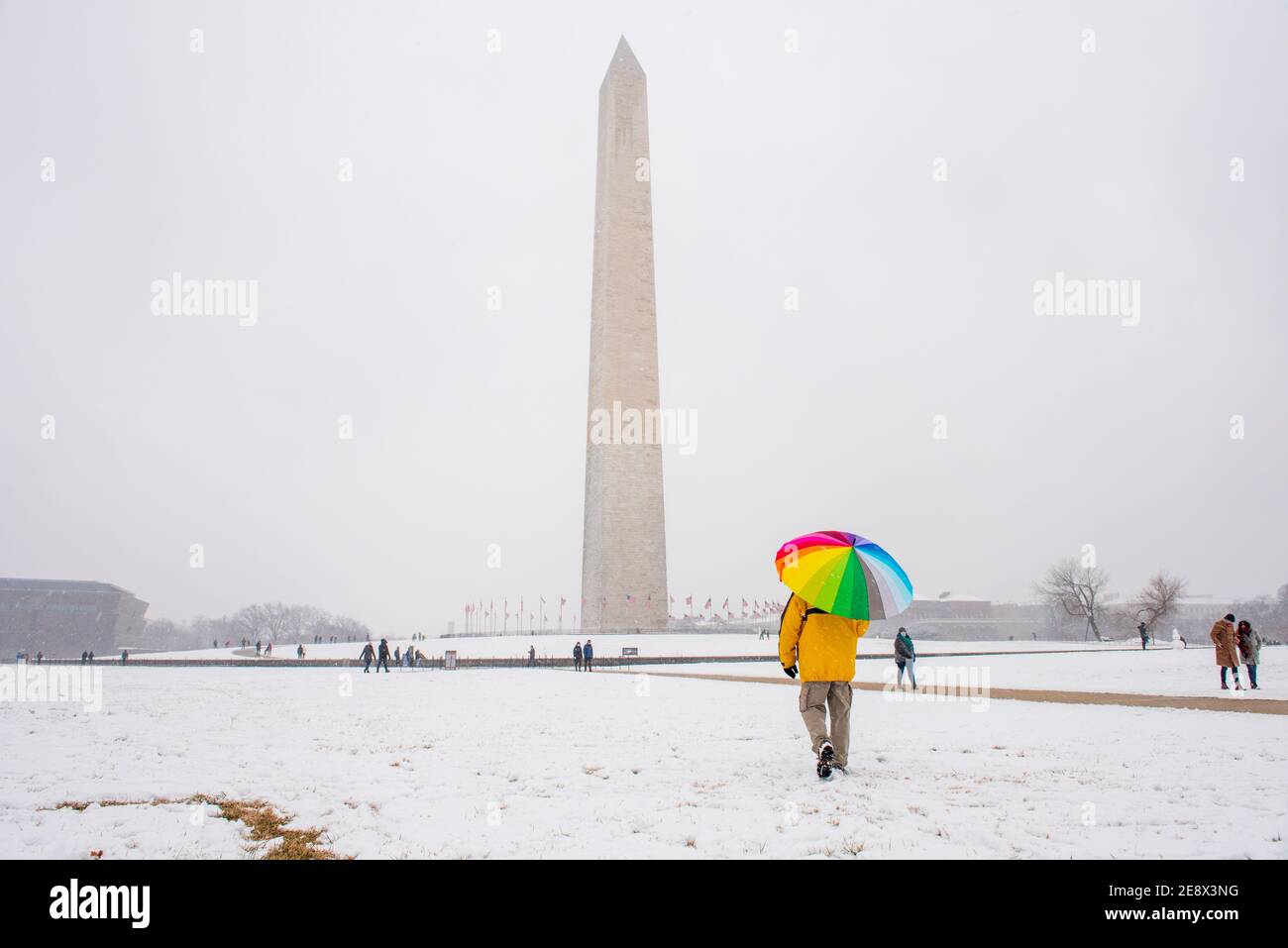Ein Mann trägt einen bunten Regenschirm während eines verschneiten Tages auf der National Mall in Washington, D.C. das Washignton Monument kann in der Ferne gesehen werden. Stockfoto