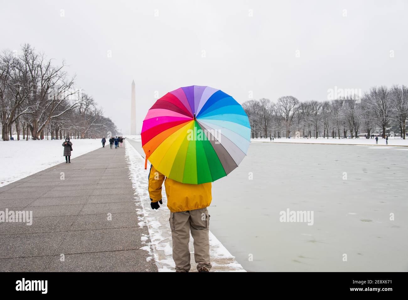 Ein Mann trägt einen bunten Regenschirm während eines verschneiten Tages auf der National Mall in Washington, D.C. das Washignton Monument kann in der Ferne gesehen werden Stockfoto