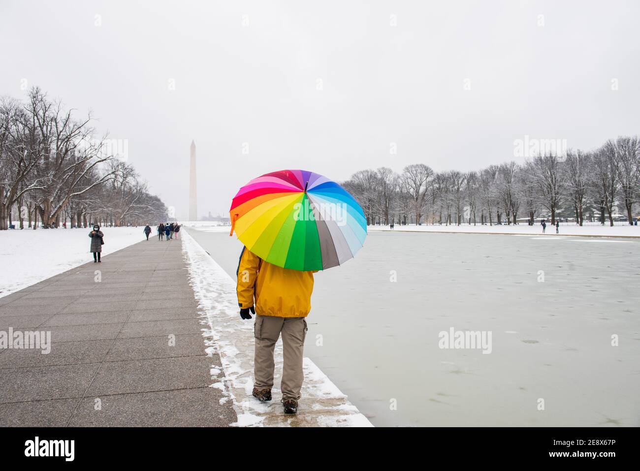 Ein Mann trägt einen bunten Regenschirm während eines verschneiten Tages auf der National Mall in Washington, D.C. das Washignton Monument kann in der Ferne gesehen werden Stockfoto