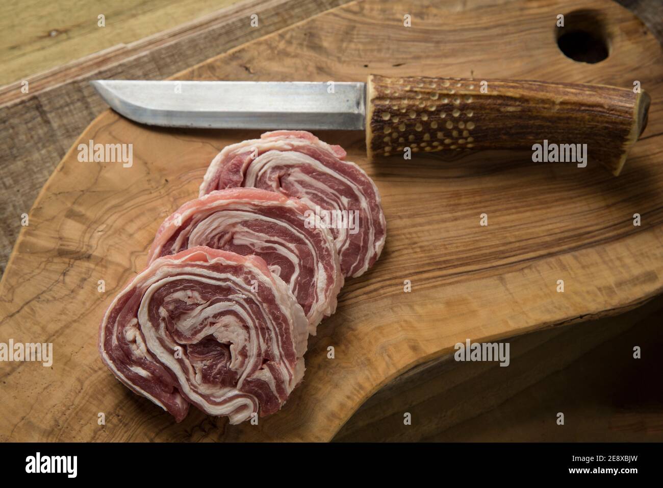 Drei Scheiben rohes, gerolltes britisches Lamm auf einem Schneidebrett aus Olivenholz mit einem hausgemachten Messer. England GB Stockfoto