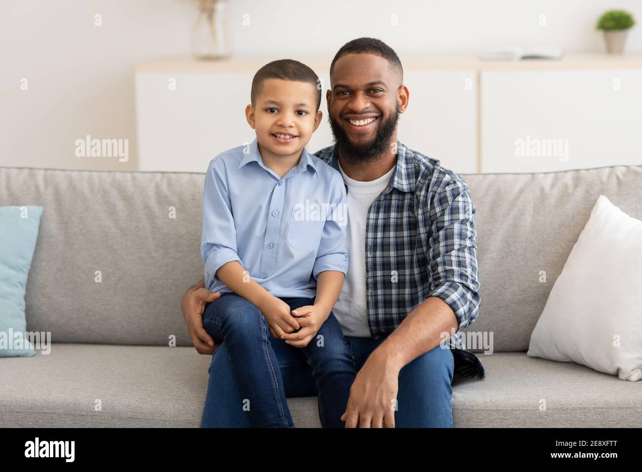 Kleiner schwarzer Junge, der auf Dads Knien sitzt und zu Hause posiert Stockfoto