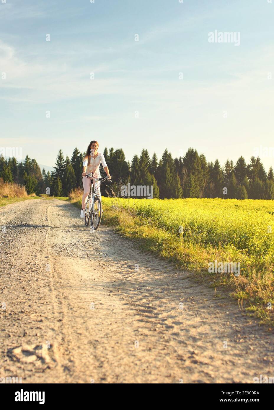 Junge Frau im Sommer leichte Kleidung fährt Fahrrad auf staubigen Straße in Richtung Kamera, Nachmittag Sonne scheint auf Felder und Wald im Hintergrund Stockfoto
