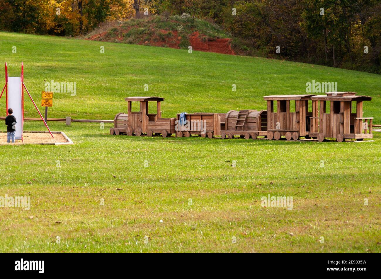 Der 4-6-jährige kaukasische Junge steht vor einer Slipper-Rutsche in einem Spielpark mit einem großen Holzzug. USA. Stockfoto Der 4-6-jährige kaukasische Junge steht vor einer Slipper-Rutsche in einem Spielpark mit einem großen Holzzug. USA. Stockfoto