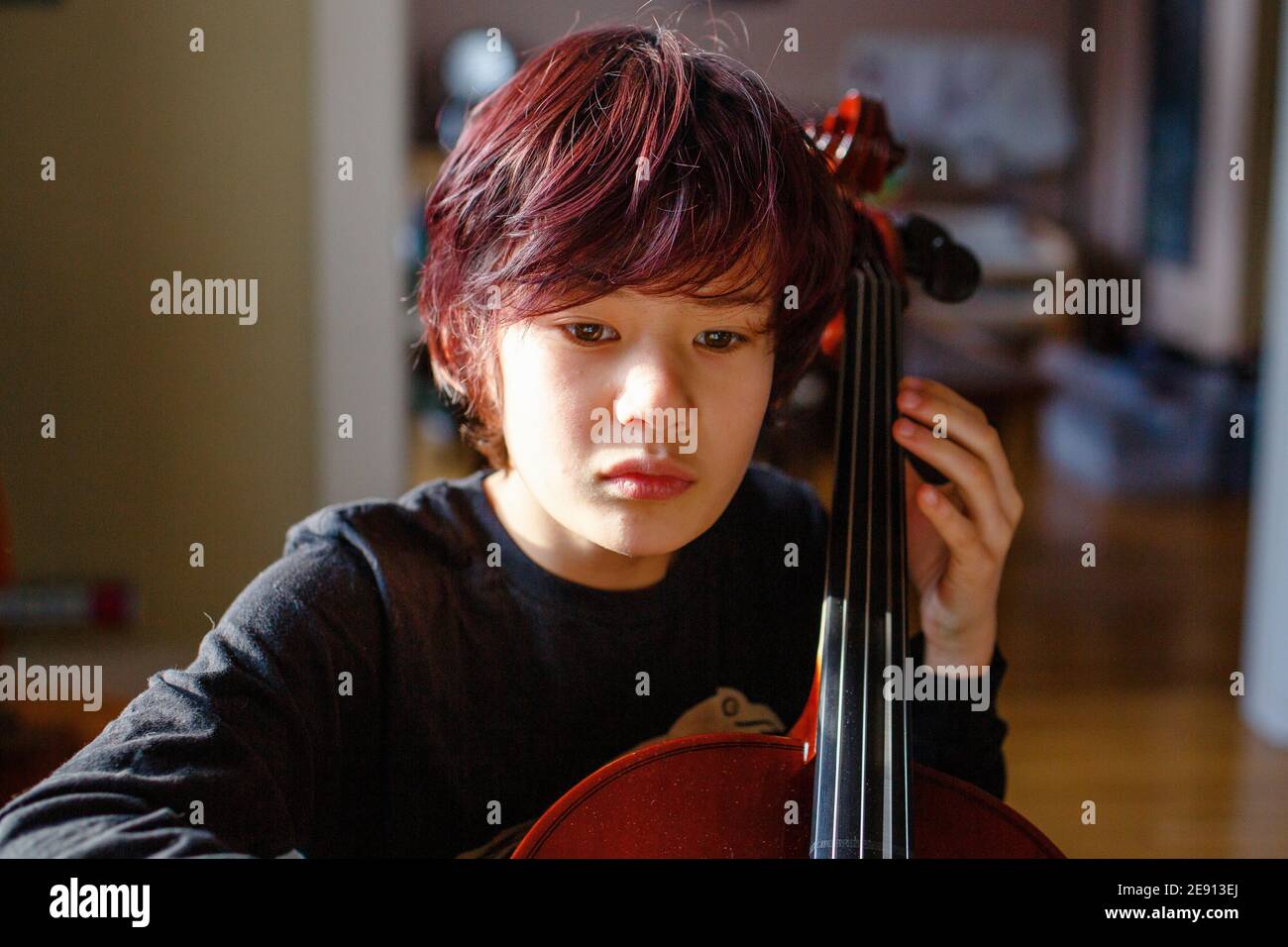 Ein Junge mit intensivem Fokus und gefärbten roten Haaren spielt Cello im Fensterlicht Stockfoto