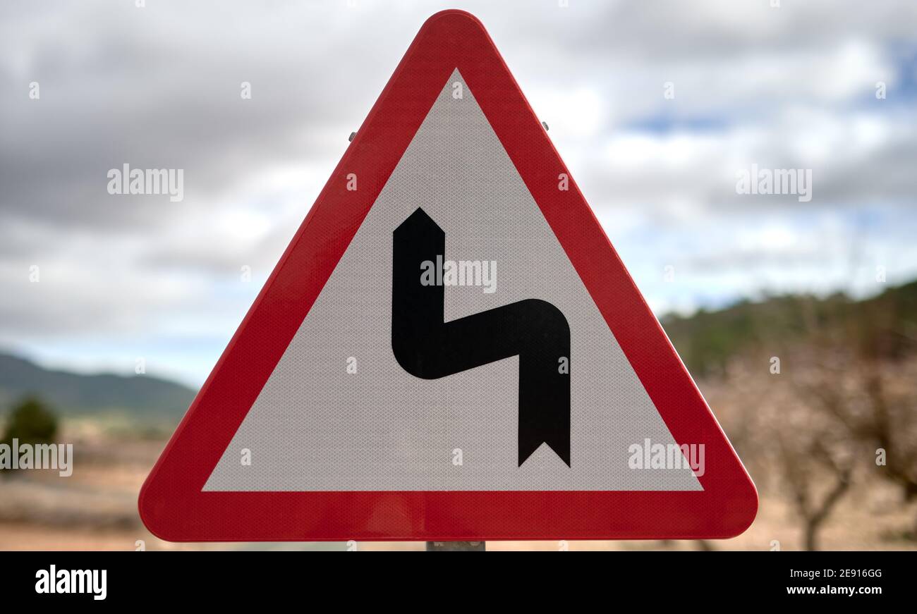 Detail eines dreieckigen Verkehrsschilds, das eine scharfe Kurve darstellt, mit rotem Rand und weißem Hintergrund Stockfoto