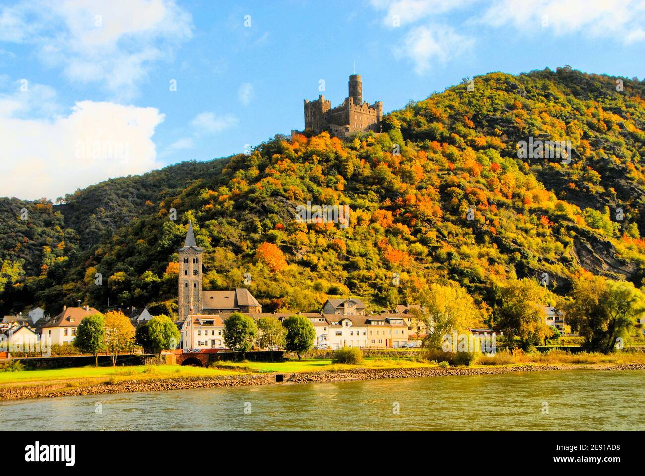 Eine Burg steht vor einem blauen Himmel mit weißen Wolken auf einem herbstlichen Hügel in der Rheinschlucht, während eine kleine Stadt unten am Rhein liegt. Stockfoto