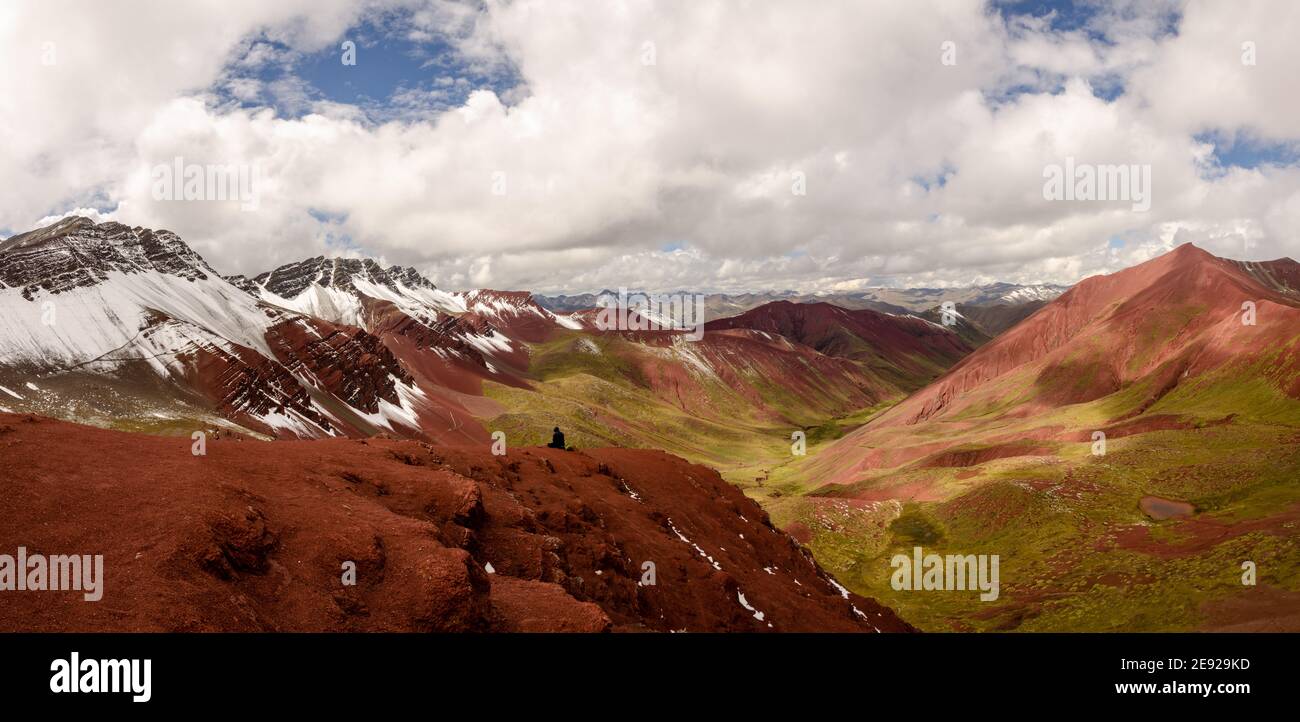 Eine Person sitzt auf einem Plateau im Red Valley, in Peru, und genießt den Blick auf das schöne rote und grüne Tal und die schneebedeckten Berge Stockfoto