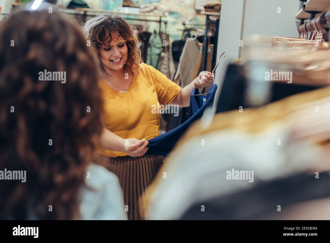 Schöne Frau hält einen Kleiderbügel mit einem neuen Kleid im Modehaus, während Verkäuferin vor sich steht. Weibliche Shopper einkaufen im Bekleidungsgeschäft. Stockfoto
