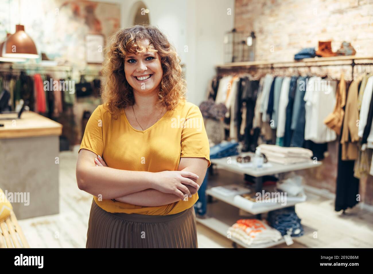 Schöne Frau, die die Arme gekreuzt hält und die Kamera anschaut, während sie im Bekleidungsgeschäft steht. Die Inhaberin der Modeboutique steht in ihrem Geschäft. Stockfoto