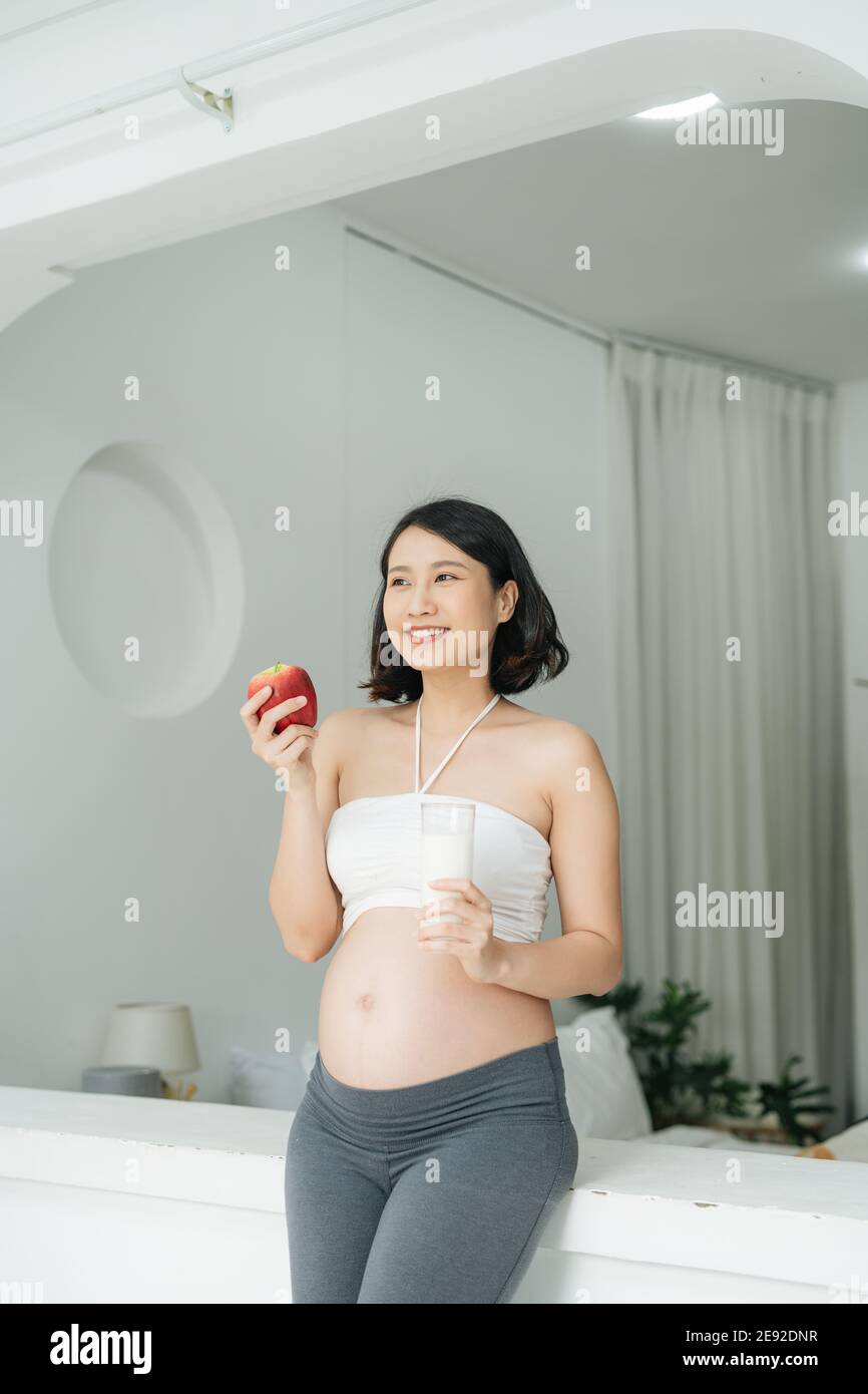 Schwangere Frau mit hübschen Magen hält roten Apfel und eine Flasche Milch, schwangere Mutter schaut nach unten Stockfoto