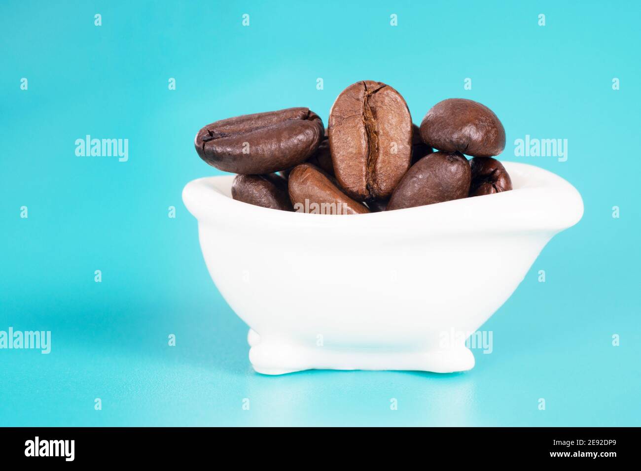 Nahaufnahme einer winzigen weißen Badewanne mit gerösteten Kaffeebohnen auf hellblauem Hintergrund. Morning Routine kreatives Konzept. Studioaufnahme, selektiver FOC Stockfoto
